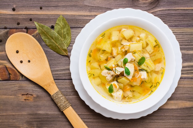 Fresh chicken broth soup with potatoes and herbs in a white bowl and wooden spoon on a wooden table. Top view