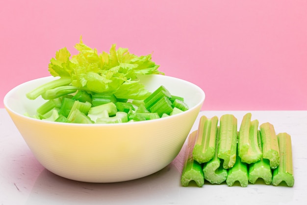 Fresh chopped celery slices in white bowl with celery sticks on white cutting board