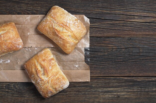 Fresh ciabatta bread on dark wooden table with paper package, top view. Space for your text