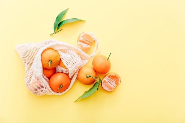 Fresh clementines with leaves in reusable string bag on yellow background, top view