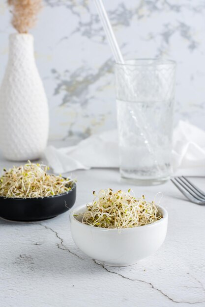 Fresh clover and alfalfa sprouts for food in a bowl and a glass of water on a light table Detox organic diet Vertical view