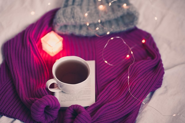 Fresh coffee in white cup staying on open book with candles on knitted sweater in bed closeup. Winter time.