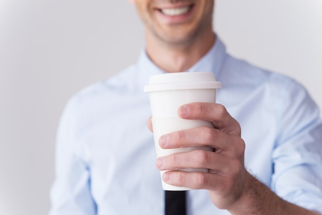 Свежий тебе кофе! Крупный план молодого человека в рубашке и галстуке, протягивающего чашку кофе и улыбающегося