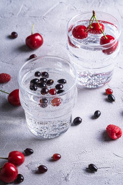 Fresh cold sparkling water drink with cherry, raspberry and currant berries in two transparent glass on stone concrete surface, summer diet beverage, angle view macro