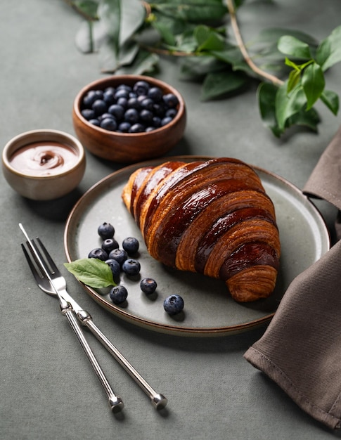 Fresh croissant with chocolate and blueberries on a dark plate on a green background with branch Delicious homemade breakfast concept