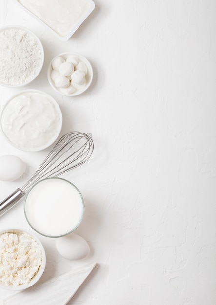 Fresh dairy products on white table. Glass of milk, bowl of flour, sour cream and cottage cheese and eggs. Steel whisk