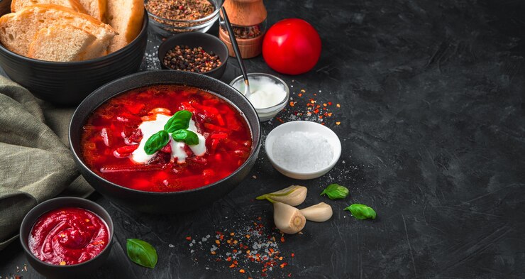 Свежий, вкусный борщ в глубокой черной тарелке, на фоне хлеба, специй, чеснока и сметаны на черном фоне. Панорама с местом для копирования.