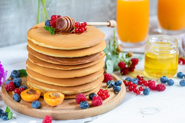 Fresh delicious pancakes with summer raspberries blueberries red currants apricot peach and mint Two glasses of orange juice honey light background Sweet nutritious food