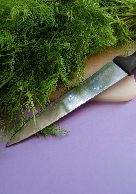 A fresh dill on a cutting board next to a knife on a purple surface.