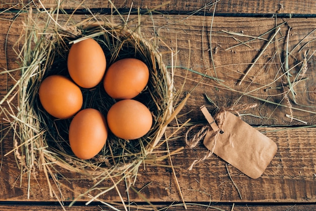 신선한 계란. 소박한 나무 탁자에 놓여 있는 건초와 갈색 종이 라벨이 있는 그릇에 있는 갈색 계란의 상위 뷰