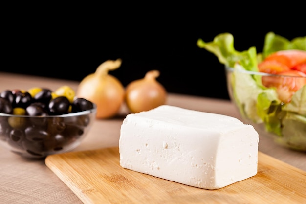 Fresh feta cheese on wooden board with a bowl of salad and olives on black background