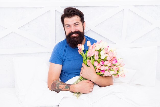 Fresh flowers surprise Flowers delivery service Birthday anniversary holiday Gift for spouse Bearded hipster in bed Spring in bedroom Man hold tulips bouquet while relaxing in bed Tender bloom