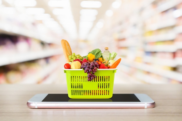 Fresh food and vegetables in shopping basket on mobile smartphone on wood table with supermarket aisle blurred background grocery online concept