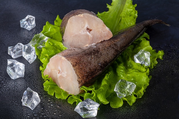 Fresh frozen hake carcass. Fish fillet with culinary ingredients, herbs, pepper, and lemon on a black surface. Copy space