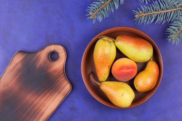 Fresh fruits for vegan closeup Ripe juicy pears and peach in a clay plate with fir branches on a blue background