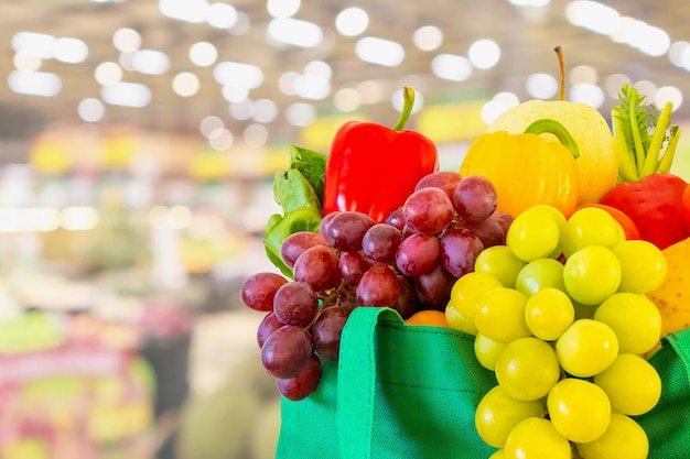 Fresh fruits and vegetables in reusable green shopping bag with supermarket grocery store blurred defocused background with bokeh light