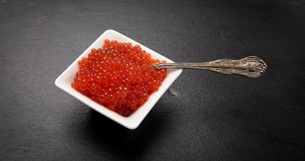 Fresh grained red chum salmon caviar in a white ceramic bowl on a black slate stone