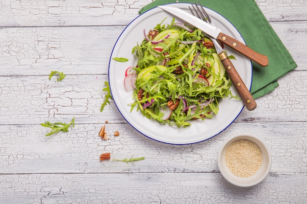 Fresh green arugula leaves on white bowl, rucola rocket salad with apple, radish, pecan nuts, onion on wooden rustic background with place for text. Top view,  healthy food, diet. Nutrition concept