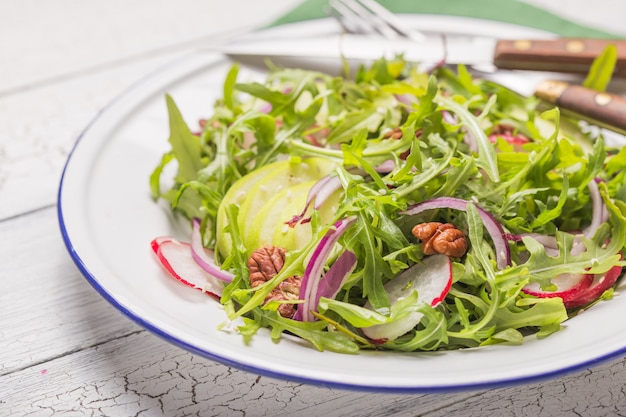 Fresh green arugula leaves on white bowl, rucola rocket salad with apple, radish, pecan nuts, onion on wooden rustic background