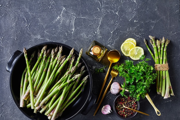 Fresh green asparagus in black baking dish with golden cutlery, lemon slices, peppercorns and bottle of olive oil on the grey concrete table