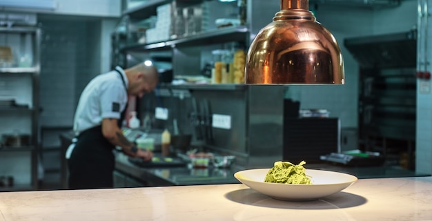 Photo fresh green salad on the plate standing on table under the light with working chef on the background