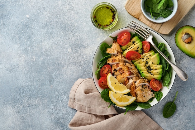 Fresh green salad with grilled chicken fillet, spinach, tomatoes, avocado, lemon and black sesame seeds with olive oil in white bowl on light slate background. Nutrition Diet Concept. Top view.