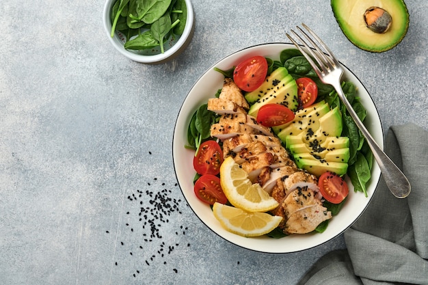 Fresh green salad with grilled chicken fillet, spinach, tomatoes, avocado, lemon and black sesame seeds with olive oil in white bowl on light slate background. Nutrition Diet Concept. Top view.