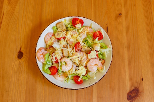 Свежий здоровый салат Цезарь с креветками, вишнями, помидорами, салатами, пармезаном, сырным соусом и крутонами на деревянном столе.