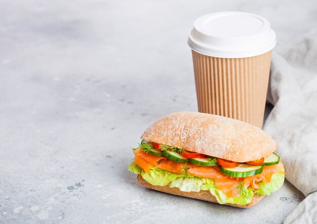 Свежий здоровый salmon сандвич с салатом и огурцом с бумажным стаканчиком кофе на белой каменной предпосылке. Закуска к завтраку.