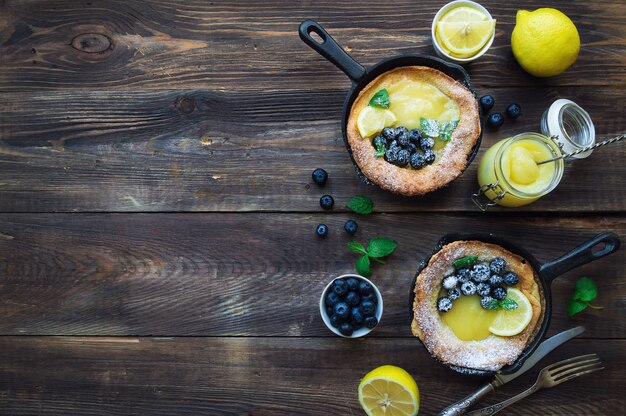 Fresh homemade Dutch Baby pancake with lemon curd and blueberries in iron skillets on rustic wooden background. Top view. Copy space area.