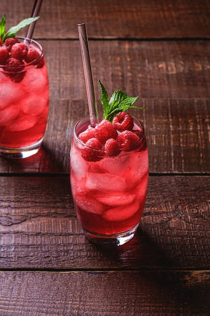 Свежий ледяной ягодный сок пить с мятой, летний малиновый лимонад в два стакана с соломкой на коричневой деревянной фоне, угол зрения селективный фокус