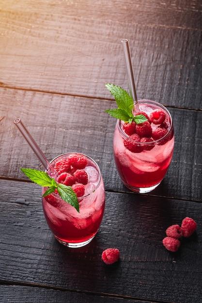 Fresh ice cold berry juice drink with mint, summer raspberry lemonade in two glass with straws on dark wooden background, angle view