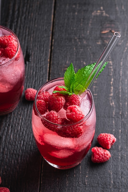 Fresh ice cold berry juice drink with mint, summer raspberry lemonade in two glass with straws on dark wooden table, angle view