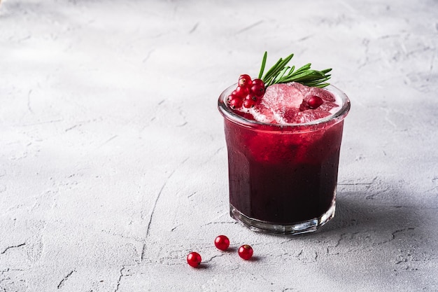 Fresh ice cold fruit cocktail in glass, refreshing summer red currant berry drink with rosemary leaf on stone concrete background, angle view copy space