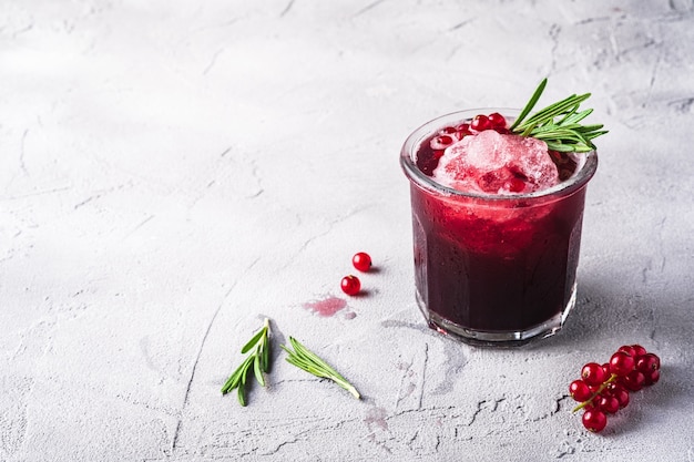 Fresh ice cold fruit cocktail in glass, refreshing summer red currant berry drink with rosemary leaf on stone concrete table, angle view copy space