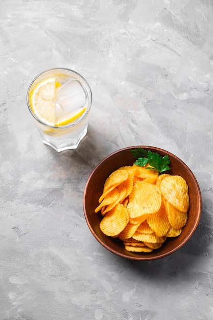 Fresh ice cold water drink with lemon near to fried corrugated golden potato chips with parsley leaf in wooden bowl on concrete background, angel view