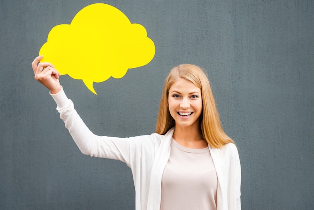 Fresh idea. Beautiful young woman holding empty speech bubble and looking at camera while standing against grey background