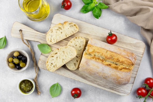 Fresh italian ciabatta bread with herbs, olive oil, black and green olives, basil leaves and pesto sauce on light gray concrete surface