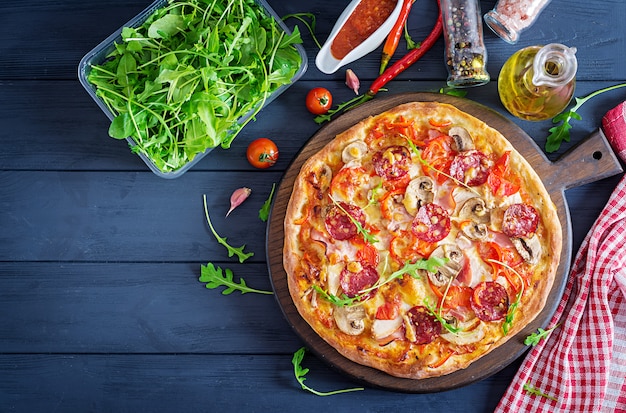 Свежая итальянская пицца с куриным филе, грибами, ветчиной, салями, помидорами, сыром на черном фоне.
