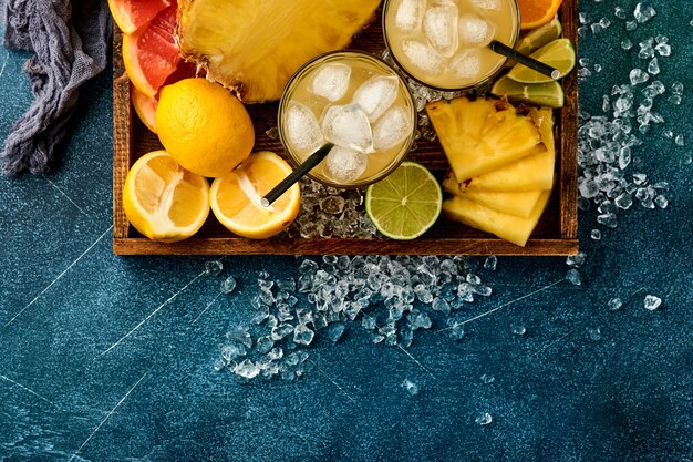 Fresh juice pineapple with ice and variety sliced ripe of color tropical fruits grapefruit, orange, lime, lemon, flat lay composition, on dark blue stone background. 