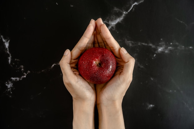 Свежее сочное красное яблоко с капельками воды на яблоке в руках в виде лотоса, держащего над черной мраморной поверхностью