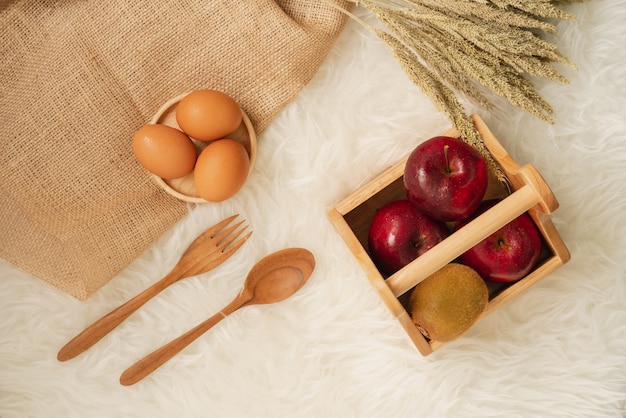 Fresh juicy red apples and kiwi fruit in wooden basket with eggs on wooden coaster and burlap cloth