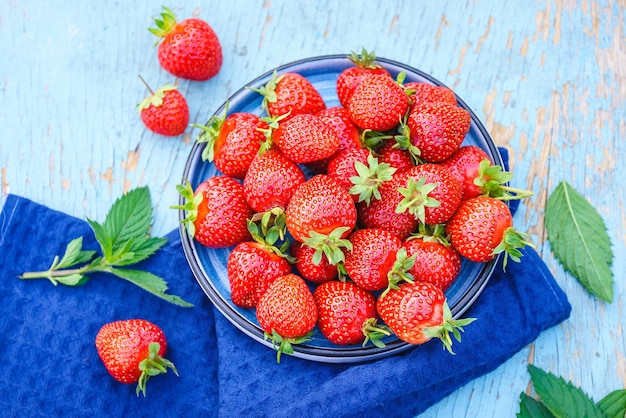 Fresh juicy strawberries in a blue plate on an old wooden rural table next to it is a dark blue napkin and mint leaves