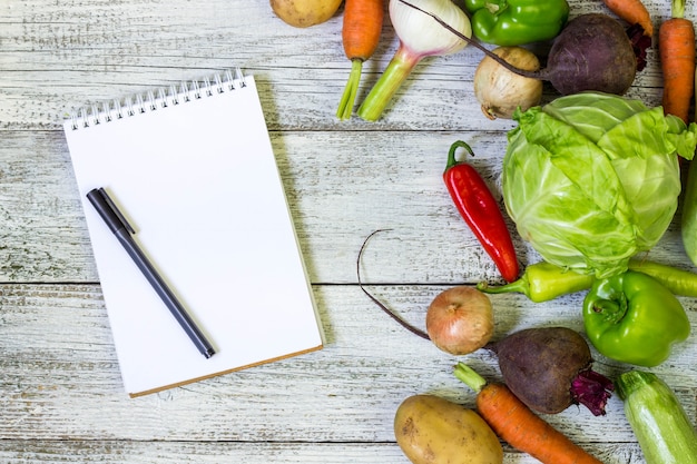 Fresh juicy vegetables border, blank white notepad   and pen on white wooden background, top view.