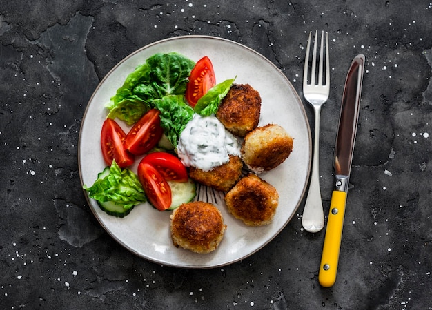 Fresh kale tomato salad and canned tuna potato fish balls with greek yogurt cilantro sauce on dark background top view