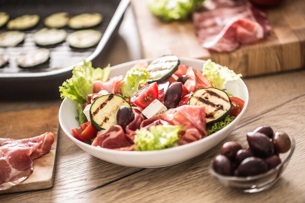 Fresh lettuce salad with grilled zucchini coppa di parma ham feta cheese olives tomatoes and olive oil.