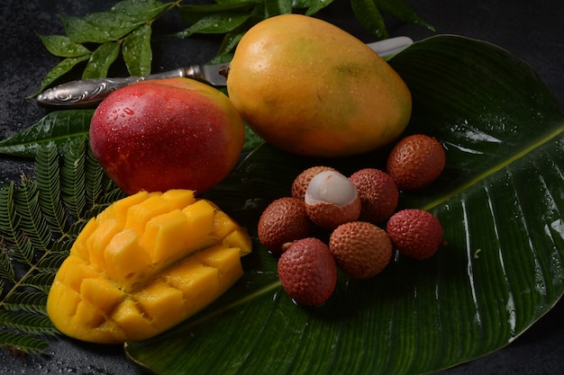 Foto frutto di mango fresco con pezzi di mango a dadini a fette su un piatto e litchi freschi e sbucciati che mostrano la pelle rossa e la polpa bianca su sfondo scuro.