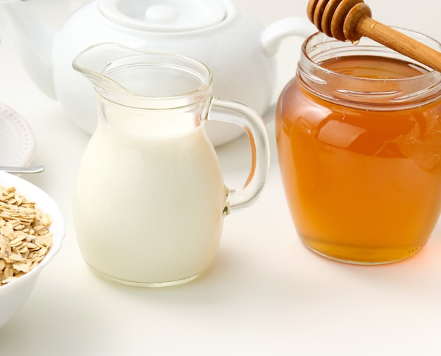 Photo fresh milk in a transparent glass decanter and a jar of honey stand on a white table, a healthy product, breakfast