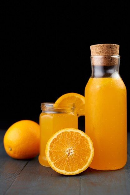 Fresh orange juice in a glass jar and bottle with fresh fruits on wooden table, black background