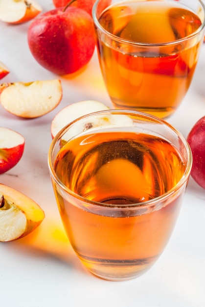Свежий органический яблочный сок фермы в стаканах с сырыми целыми и нарезанными красными яблоками, на белом мраморном столе, copyspace